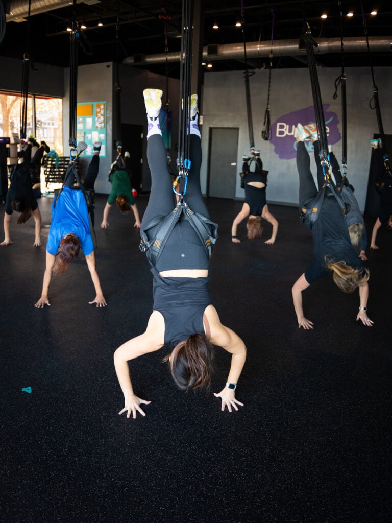 Group bungee fitness class hanging upside down in harnesses with hands on the floor