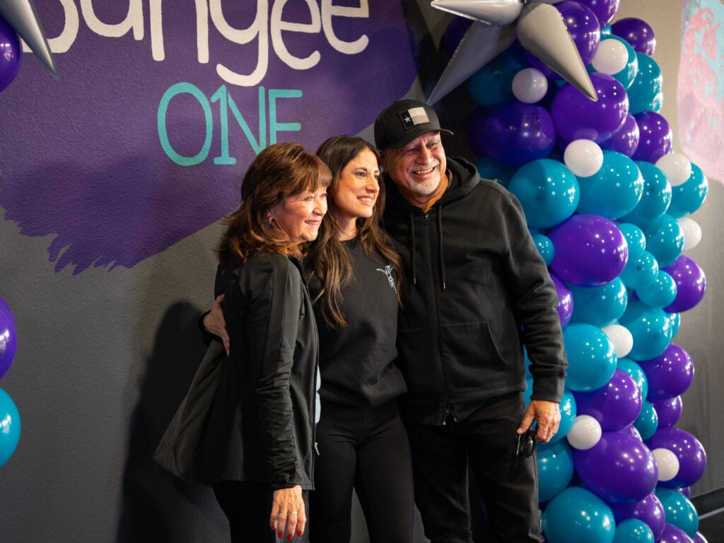 Three people hugging and smiling beside balloons at the BungeeONE grand opening